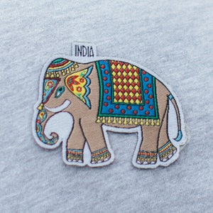 Può includere: Una toppa ricamata colorata di un elefante con la parola "INDIA" sopra. L'elefante è decorato con una coperta a motivi blu, rosso, giallo e bianco.