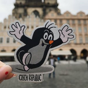 Könnte beinhalten: Ein Cartoon-Maulwurf, der ein graues und schwarzes Outfit mit einer orangefarbenen Nase trägt, winkt mit beiden Händen. Der Maulwurf steht auf einem grauen Surfbrett mit dem aufgedruckten Text "Tschechische Republik". Der Maulwurf befindet sich vor einem verschwommenen Hintergrund einer Kirche mit zwei Türmen.