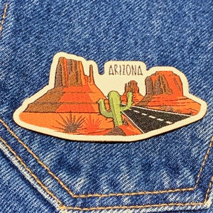 Può includere: Un cerotto in pelle marrone con un'immagine ricamata di un paesaggio desertico con formazioni rocciose rosse, un cactus e una strada. Il testo "ARIZONA" è ricamato in bianco sul cerotto.