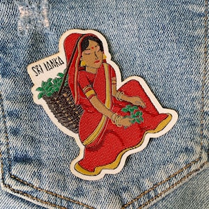 Może przedstawiać: Naszywka haftowana przedstawiająca kobietę w czerwono-złotym sari trzymającą kosz z zielonymi liśćmi. Naszywka jest opisana napisem "Sri Lanka" białymi literami.