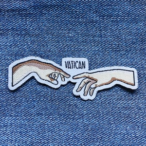 Peut inclure: Un patch brodé représentant deux mains se touchant, en blanc et marron, avec le mot "VATICAN" au-dessus.