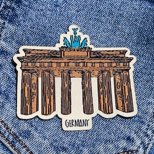 Könnte beinhalten: Ein braunes und weißes gesticktes Patch der Brandenburger Tor in Berlin, Deutschland. Das Patch hat einen blauen und weißen Adler oben und das Wort "GERMANY" darunter.