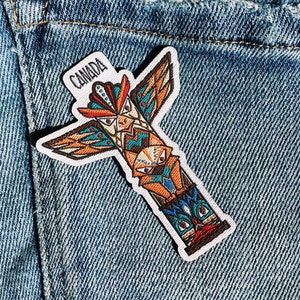 Puede incluir: Un parche bordado colorido que presenta un tótem con un diseño de pájaro y la palabra "CANADA" en letras blancas. El parche está cosido en un bolsillo de mezclilla azul.