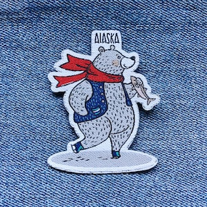 May include: A gray bear wearing a blue vest and a red scarf, holding a fish in its right paw. The bear is standing on a white background with the word "Alaska" written above it.