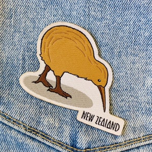 May include: An embroidered patch featuring a brown kiwi bird with a white background and the text "NEW ZEALAND" on a white label.