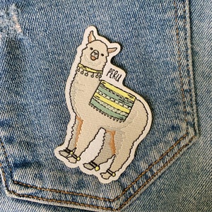 May include: A white llama with a yellow and green striped blanket and the word "PERU" on its side, embroidered on a blue denim background.