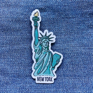 Puede incluir: Un parche bordado azul y verde que representa la Estatua de la Libertad sosteniendo una antorcha, con el texto "NEW YORK" debajo.