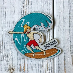 以下が含まれることがあります： 「INDONESIA」と書いてあるサーフボードに乗った、漫画のサーファーが波に乗っている。サーファーは赤いショートパンツを履いていて、金髪である。波は青と緑である。