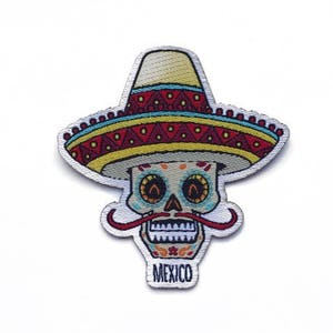 May include: A colourful embroidered patch of a sugar skull wearing a sombrero. The skull has a white face with black eyes, a red nose, and a red moustache. The sombrero is yellow, red, and blue with a white trim. The text "MEXICO" is printed below the skull.