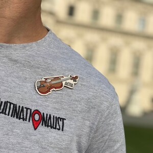May include: A gray t-shirt with the text "MULTINATIONAUST" and a red pin. A brown violin with the text "Austria" is pinned to the shirt.