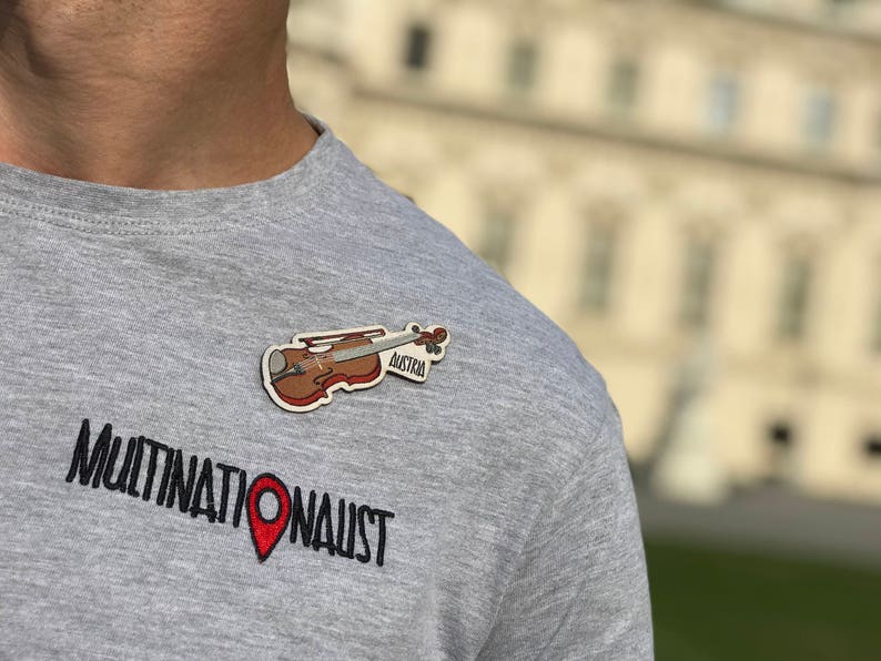 May include: A gray t-shirt with the text "MULTINATIONAUST" and a red pin. A brown violin with the text "Austria" is pinned to the shirt.