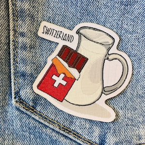 May include: A white embroidered patch with a pitcher of milk, a chocolate bar, and a Swiss flag. The text "Switzerland" is embroidered on the patch.