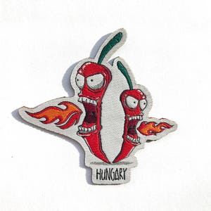 Puede incluir: Parche bordado con dos pimientos rojos de dibujos animados con llamas y bocas abiertas. El parche tiene un borde gris y la palabra "HUNGARY" en la parte inferior.