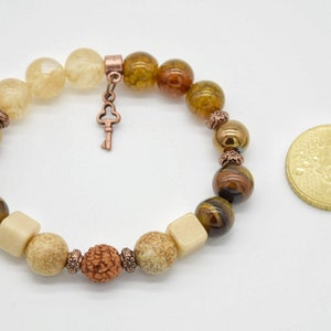 May include: A brown, beige, and gold beaded bracelet with a key charm. The bracelet is made with tiger's eye, agate, and other natural stones.