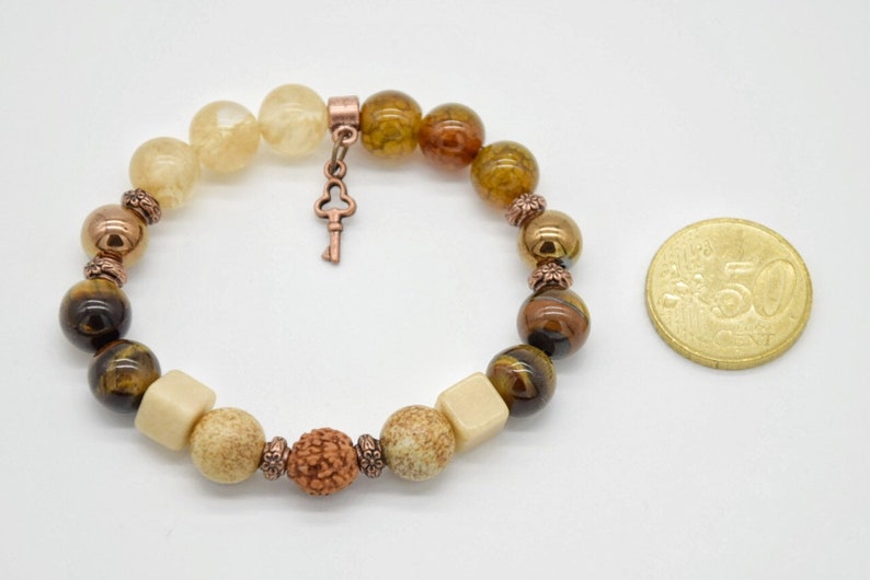 May include: A brown, beige, and gold beaded bracelet with a key charm. The bracelet is made with tiger's eye, agate, and other natural stones.