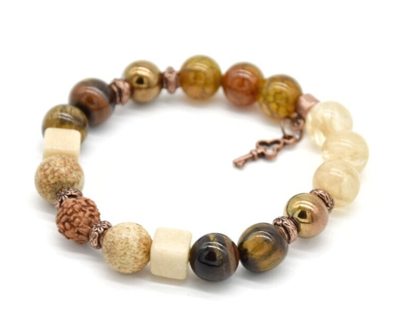 May include: A brown, beige, and gold beaded bracelet with a copper key charm. The bracelet features tiger's eye, wood, and other natural stones.