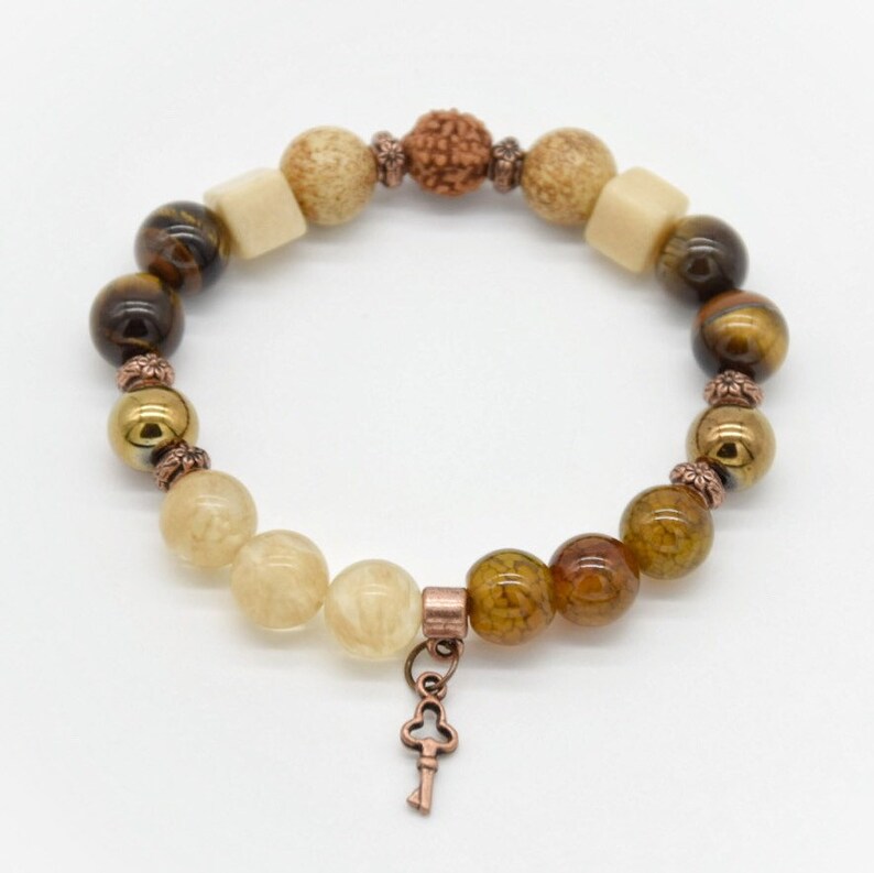 May include: A brown, beige, and gold beaded bracelet with a key charm. The bracelet features various stones, including tiger's eye, agate, and jasper.
