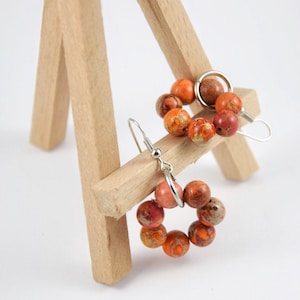 May include: A pair of orange and brown jasper earrings with silver hooks. The earrings are in the shape of a circle and are made of small beads.
