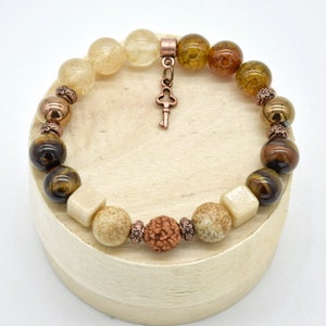 May include: A brown, beige, and gold beaded bracelet with a key charm. The bracelet is made with various stones, including tiger's eye, agate, and quartz.