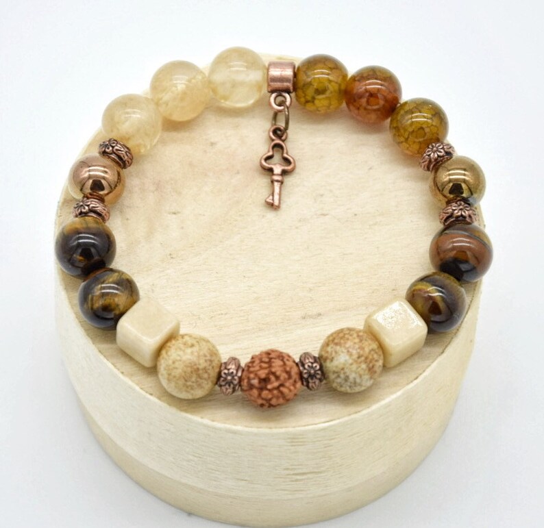 May include: A brown, beige, and gold beaded bracelet with a key charm. The bracelet is made with various stones, including tiger's eye, agate, and quartz.