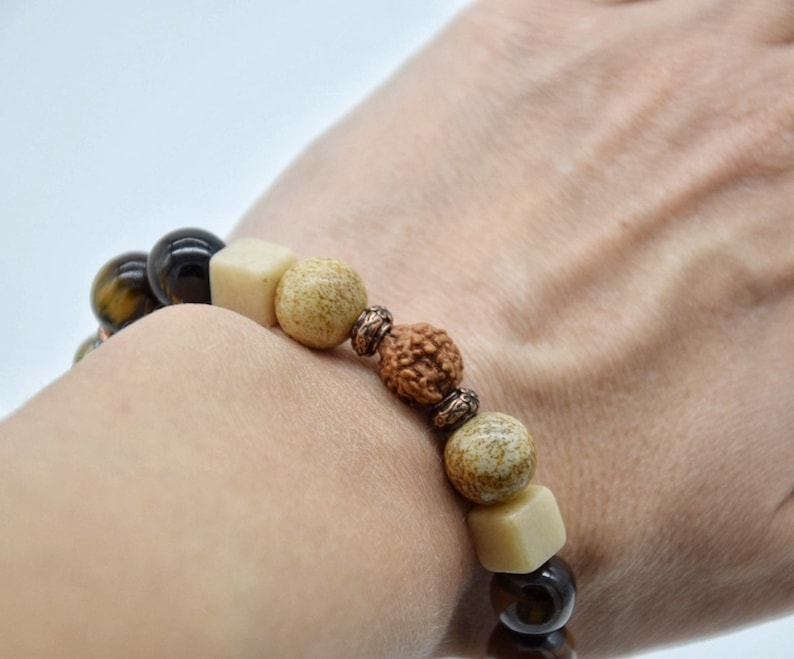 May include: A brown, black, and cream beaded bracelet with a copper accent. The bracelet is made with tiger's eye, wood, and rudraksha beads.