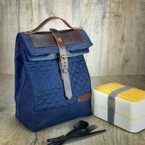 May include: A blue quilted lunch bag with a brown leather strap and buckle. The bag is sitting on a wooden surface next to a white and wood two-tiered lunch box and a set of black utensils.