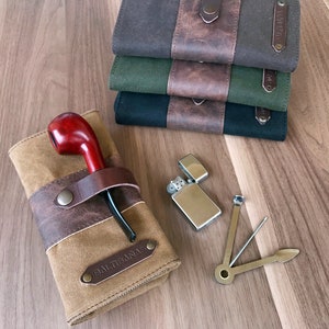 Puede incluir: Una bolsa de tabaco de lona marrón con una correa de cuero y una pipa de madera roja. La bolsa tiene el texto "BALTRAMA" impreso. También se muestra un encendedor plateado y un tampón de pipa multiherramienta.