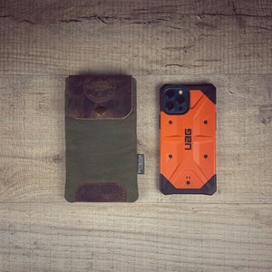 May include: An olive green canvas phone case with a brown leather flap and accents, next to an orange and black phone case. The canvas case has a leather patch with a logo and the text "BALTIBASAS". The orange case has the text "UAG".