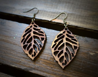 Puede incluir: Un par de pendientes de madera con forma de hoja con un diseño delicado e intrincado. Los pendientes son marrones con una veta de madera clara y tienen un gancho de color bronce.
