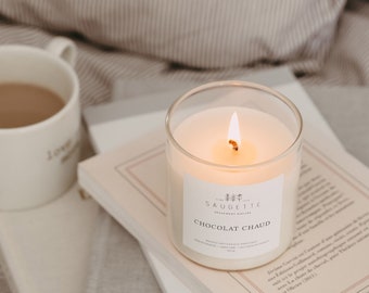 Può includere: Una tazza di ceramica bianca piena di caffè si trova su una pila di libri accanto a una candela accesa in un barattolo di vetro trasparente. La candela è etichettata "La Saugetie" e "Chocolat Chaud".