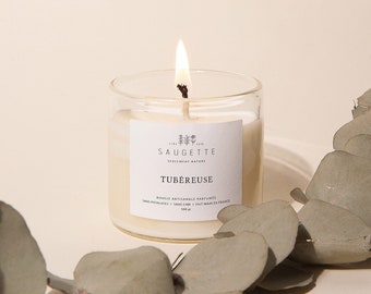 Può includere: Una candela bianca in un barattolo di vetro trasparente con un'etichetta bianca che recita "The Parisian Saugette, Soywax Candle, Tubereuse". La candela è accesa e circondata da foglie di eucalipto.