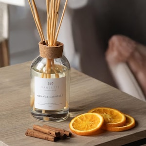 May include: A clear glass diffuser bottle with a cork top and reeds, labeled "SAUGETTE ORANGE CANNELLE." Beside it are orange slices and cinnamon sticks. The diffuser sits on a wooden surface.