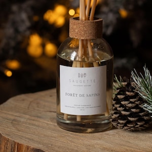 Op de afbeelding: Een glazen diffuserfles met houten stokjes en een kurken stop. Het etiket luidt "SAUGETTE FORÊT DE SAPINS". Een dennenappel en een takje den liggen naast de fles op een houten oppervlak. De achtergrond heeft vage lichten.