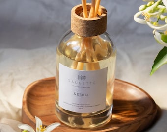 May include: A clear glass diffuser bottle with a cork top and wooden reeds, labeled "NÉROLI" by SAUGETTE. The bottle sits on a wooden tray, with white flowers and a soft background.