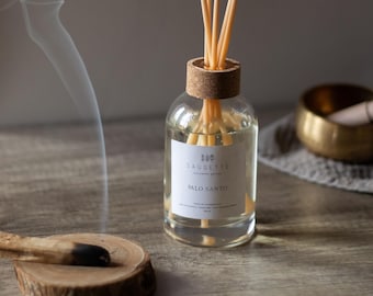 May include: A clear glass diffuser bottle with a cork top and wooden reeds. The label reads "SAUGETTE PALO SANTO". A piece of burning Palo Santo wood sits on a wooden disc, with smoke rising. A gold bowl is in the background.