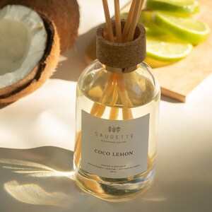 Könnte beinhalten: Ein Glas-Diffusor mit Korkverschluss und Holzstäbchen, beschriftet mit "Coco Lemon" von Saugette. Die Flasche steht neben einer halbierten Kokosnuss und Limettenscheiben auf einem Holzbrett. Der Diffusor enthält 200 ml Flüssigkeit.