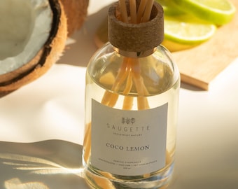 May include: A glass diffuser bottle with a cork top and wooden reeds, labeled "Coco Lemon" by Saugette. The bottle sits next to a halved coconut and lime slices on a wooden board. The diffuser contains 200 ml of liquid.