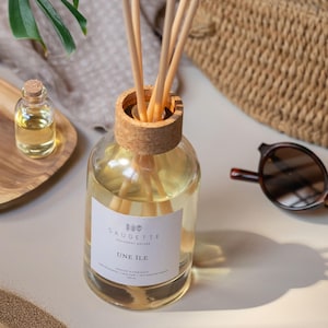 Peut inclure: Un diffuseur en verre transparent avec un bouchon en liège et des bâtonnets en bois. La bouteille contient un liquide jaune et porte l'étiquette "UNE ILE". Une petite bouteille en verre avec un bouchon en liège et des lunettes de soleil sont également visibles.