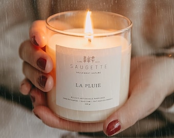 May include: A burning candle in a clear glass jar is held. The candle has a white label with the text "SAUGETTE" and "LA PLUIE". The candle is labeled as a 200g artisanal scented candle made in France.