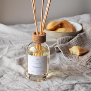 Op de afbeelding: Een heldere glazen diffuserfles met houten stokjes, een kurken dop en een label met de tekst "Madeleine". De fles staat op een linnen oppervlak, met een kom met madeleines en een servet op de achtergrond. Thema: huisgeur.