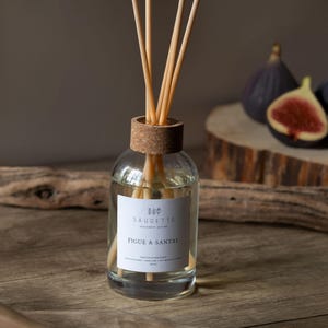 May include: A clear glass diffuser bottle with wooden reeds, a cork top, and a label that reads "Figue & Santal." The bottle sits on a wooden surface, with figs in the background. The reeds are light brown.