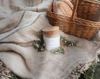 Può includere: Una candela bianca con un tappo di sughero e un'etichetta che recita "La Savonnerie - Herbe Fraiche" è appoggiata su una coperta di lino beige con un bordo a frange. La candela è circondata da piccoli fiori bianchi. Un cesto di vimini con del pane è sullo sfondo.