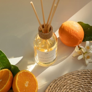 May include: A glass diffuser bottle with wooden reeds, labeled "Fleur d'Oranger", sits next to fresh oranges and orange blossoms. The bottle has a cork top. A woven coaster and green leaves complete the arrangement.