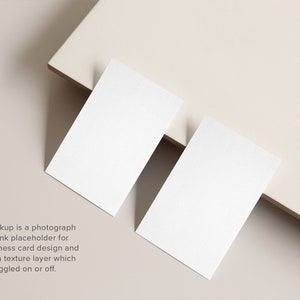 May include: Two blank white business cards on a light beige surface. The cards are positioned at an angle, with one card partially overlapping the other.