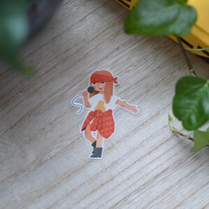 Op de afbeelding: Een sticker met een cartoonillustratie van een persoon die in een microfoon zingt. De persoon draagt een wit t-shirt, een rode geruite rok, een rode hoofdband en zwarte laarzen. De microfoon heeft een blauw snoer.