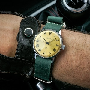 May include: A gold-faced wristwatch with black Roman numeral hour markers and a green leather strap. The watch face has the word "Poljot" printed on it.
