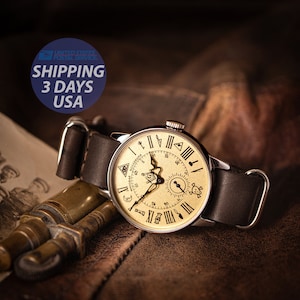 Puede incluir: Un reloj de pulsera de estilo vintage con una esfera dorada y una correa de cuero negro. La esfera del reloj presenta n&uacute;meros romanos, una br&uacute;jula y un dise&ntilde;o de calavera y huesos cruzados. El reloj est&aacute; sobre una superficie de cuero marr&oacute;n.