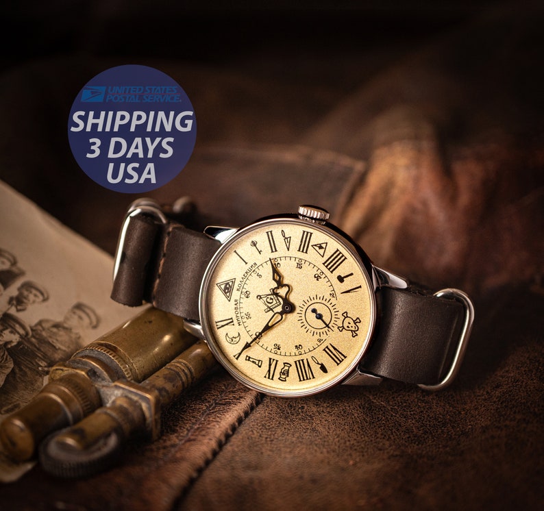 Puede incluir: Un reloj de pulsera de estilo vintage con una esfera dorada y una correa de cuero negro. La esfera del reloj presenta n&uacute;meros romanos, una br&uacute;jula y un dise&ntilde;o de calavera y huesos cruzados. El reloj est&aacute; sobre una superficie de cuero marr&oacute;n.