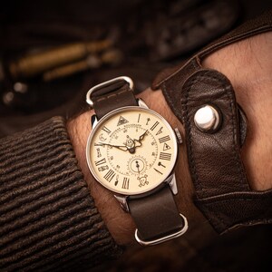 Puede incluir: Un reloj de pulsera de cuero marr&oacute;n con una esfera de color crema y marcadores de hora con n&uacute;meros romanos negros. La esfera del reloj presenta una br&uacute;jula, una calavera y huesos cruzados y un s&iacute;mbolo mas&oacute;nico.