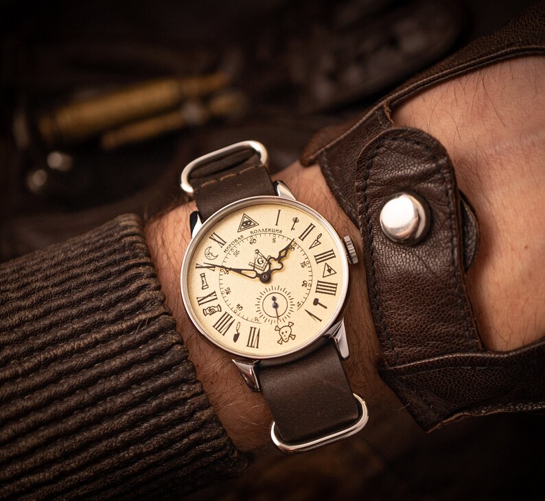 Puede incluir: Un reloj de pulsera de cuero marr&oacute;n con una esfera de color crema y marcadores de hora con n&uacute;meros romanos negros. La esfera del reloj presenta una br&uacute;jula, una calavera y huesos cruzados y un s&iacute;mbolo mas&oacute;nico.